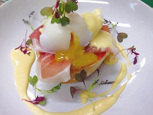 Poached egg on top of Parma ham with cream coloured Hollandaise sauce around on a white plate.