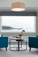 Low teal blue armchairs with a nest of round table between looking out of a window to the sea beyond.